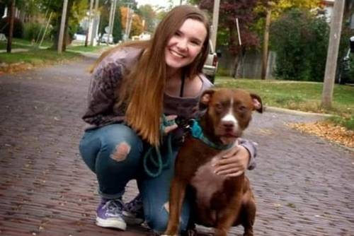 A student kneels in the road next to a dog and smiles for a photo.