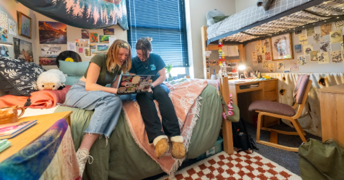 Two students sitting in their dorm room laughing at something on a laptop.