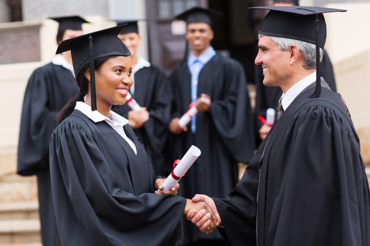 A smiling college administrator shakes hands with a graduating student wearing a cap and gown and holding a diploma.