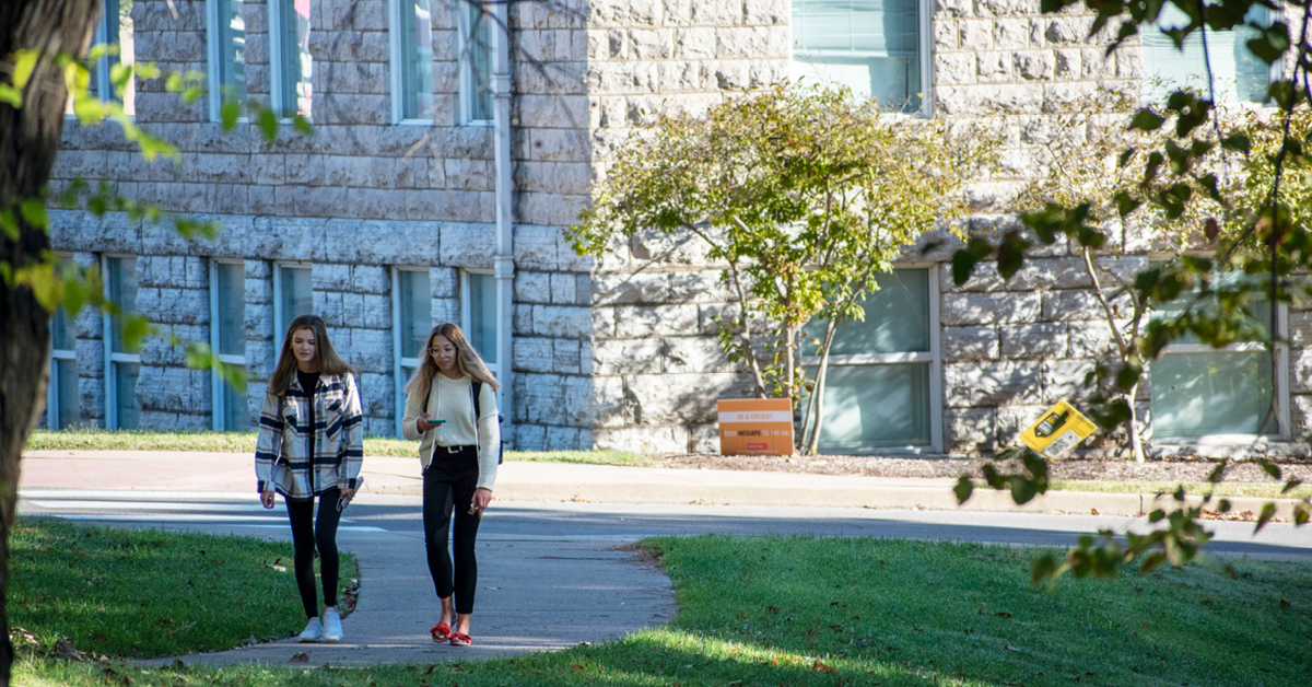 Two students walking down the sidewalk in front of Academic Hall.