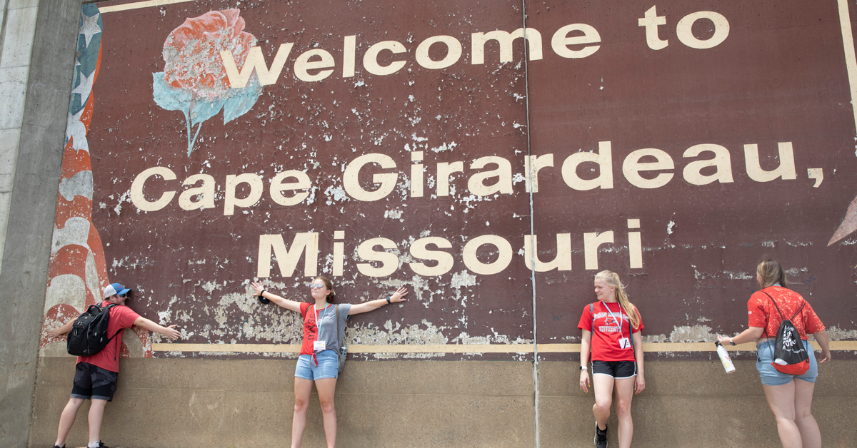 Four students in summer clothing pose in front of a river wall mural. The mural reads Welcome to Cape Girardeau Missouri and features an American flag and a rose.