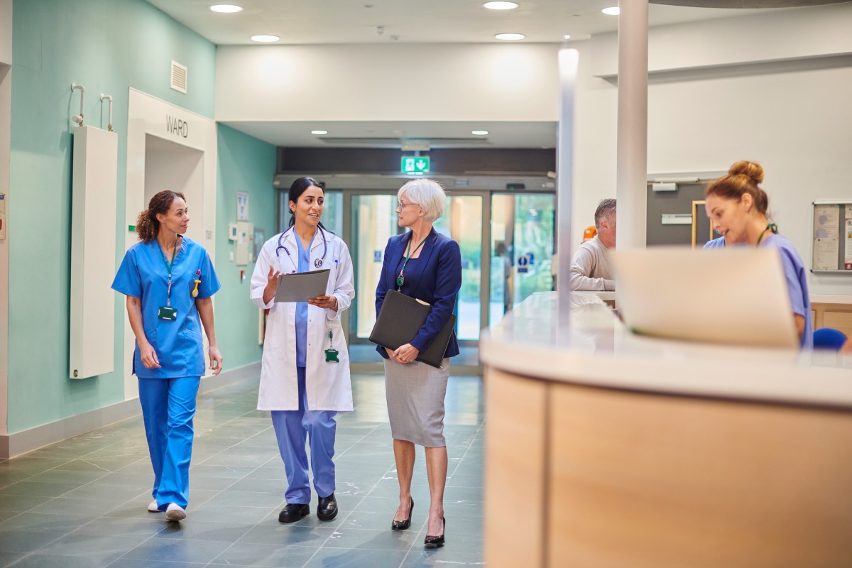 A healthcare manager talks with clinicians in a hospital corridor.