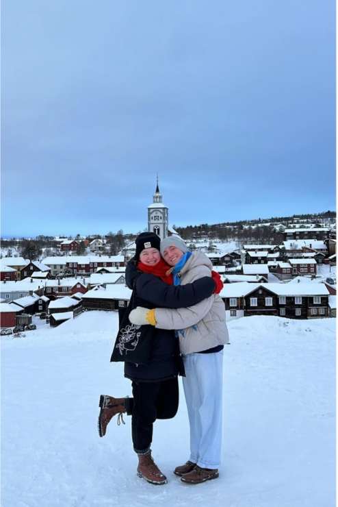 Megan in the snow on a hill with her friend. Behind them is a small city in northern Norway