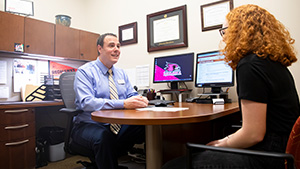 A SEMO student with Red Hair talks to their academic advisor, a person with a blue shirt and tie, as well as brown hair.