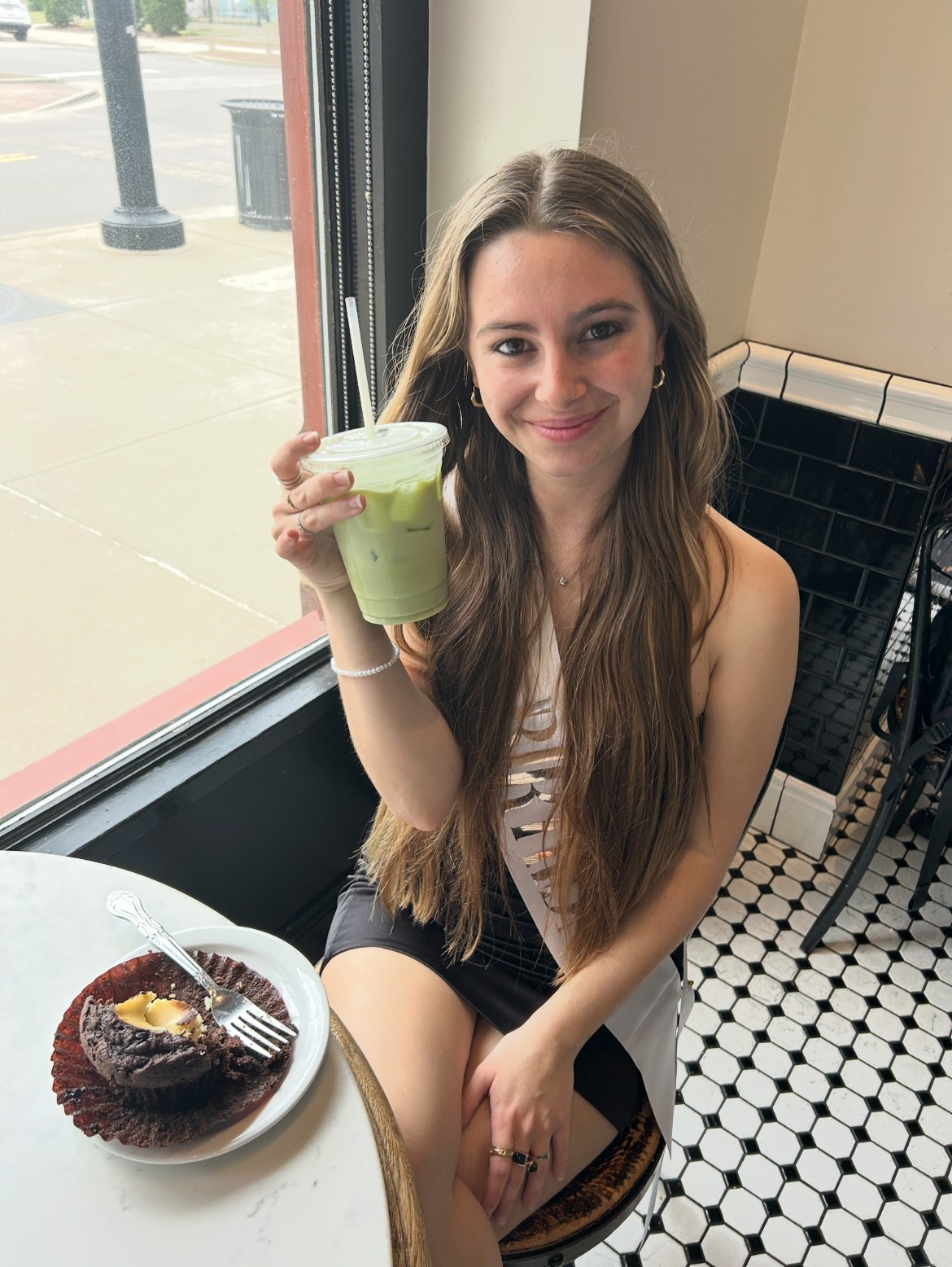 Girl at a coffee shop wearing a black dress with a birthday sash with a matcha latte and a muffin.