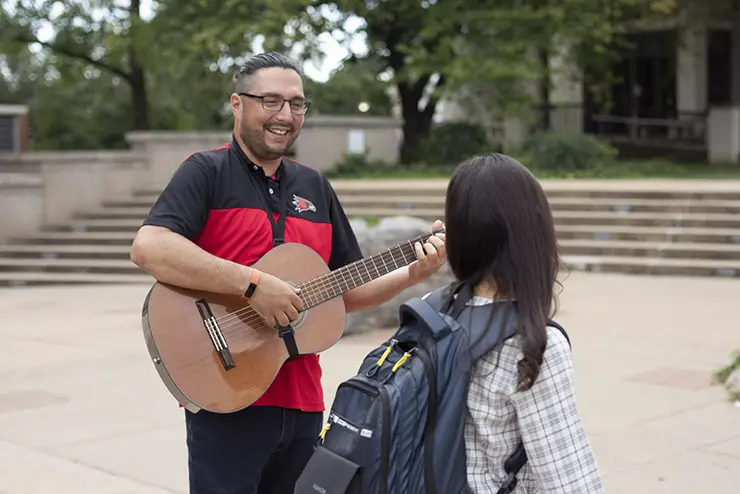 a Hispanic SEMO student plays guitar on the patio in front of Kent Library for passing students