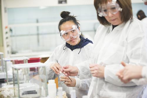 A chemistry student wearing safety goggles and a white lab coat looks up at their professor while working in the Magill chemistry lab.