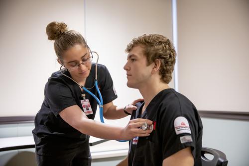 A nursing student wears a stethoscope and places the end on another nursing student to listen to their heart.