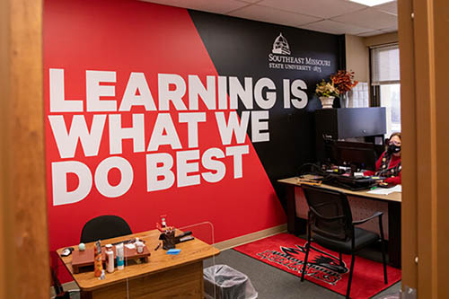 the office at the Poplar Bluff delivery location with a large wall graphic reading 'learning is what we do best' and a small desk oppsite the door with a larger, occupied desk to the right of the door