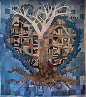 a piece of artwork created like a quilt, with patches of fibers sewn together to depict a tree with a rough heart shap behind in tans blacks and blues