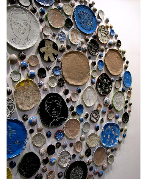 A display of different sized plates hanging on a white wall. The plates vary in color from white, to brown, to black to blue. Some have faces drawn on them while others have animals like bats and bears. 