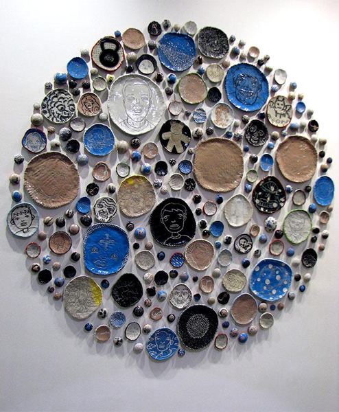 A display of different sized plates hanging on a white wall. The plates vary in color from white, to brown, to black to blue. Some have faces drawn on them while others have animals like bats and bears. 