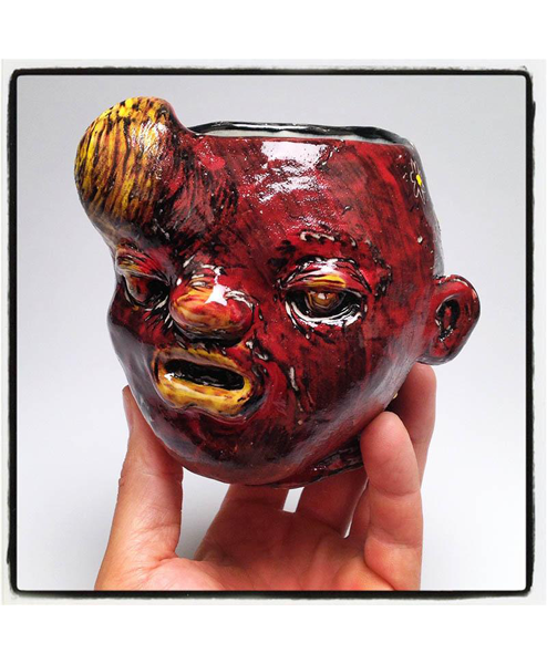 A hand holding a red ceramic mug with a face carved into it. 