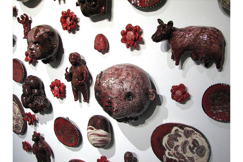 A close up of a display of ceramic pieces hanging on a wall. The pieces include heads that vary in color from red to brown. There are several animals including a cow, a pig, and a bear. The display includes several flower shaped pieces. 