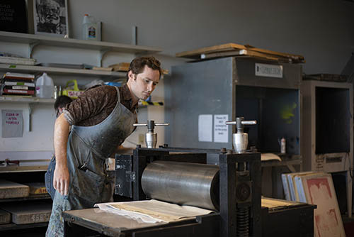 an art and design printmaking student pulls a print through the roller