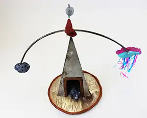 a sculpture featuring a three sided pyramid-shaped structure with a curved bar balanced on the top, one end of the bar has a meteorite and the other a cloud with streamers