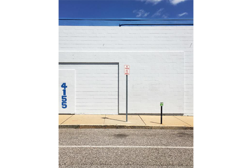 A no parking street sign in front of a white wall with a bright blue sky above it and a grey street in the foreground 