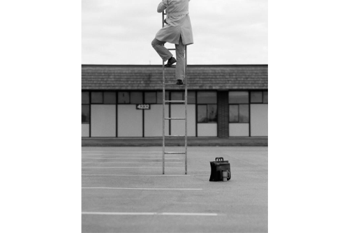 A man climbs a ladder in the middle of a parking lot with his briefcase and coffee cup on the ground and a building behind him 