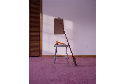 A gray stool sits in the middle of a room with mauve carpet. A broom leans against the stool with an old fashioned bell and book on top of the stool 