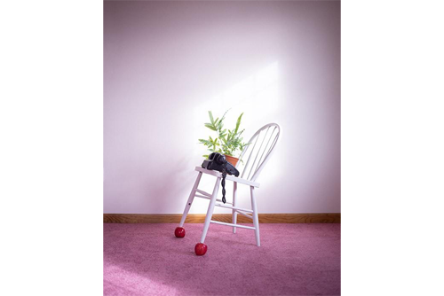 A white chair sits in a room with a white wall behind it and mauve carpet. The chair is leaning back on two legs with apples underneath the front legs. A potted plant and a telephone sit on the chair. 