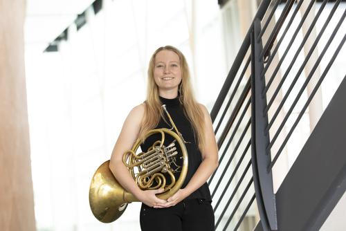 victoria wearing a black dress standing in front of a well lit window holding her french horn and smiling at the camera