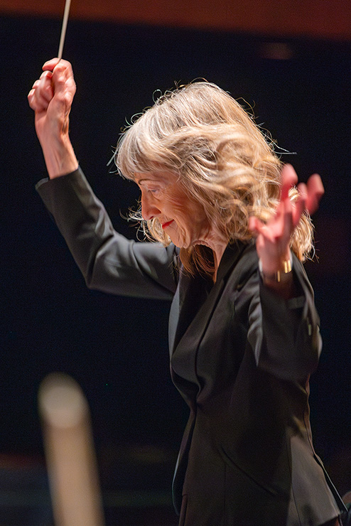During the recital, the conductor is moving the baton in the air, while holding her other hand out near her shoulder.