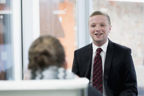 A Master of Business Administration student, in a suit and tie, smiles while giving a presentation.