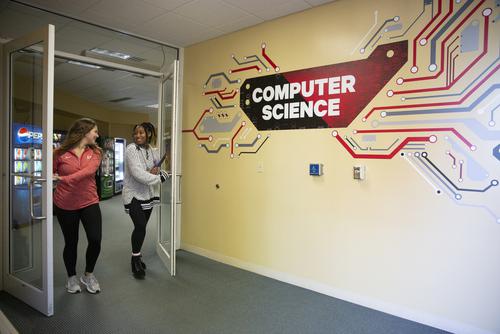 Students smile while walking along a hall featuring a “computer science” graphic on the wall.