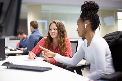two students sitting together in a computer lab, one is taking notes on a notepad and the other is pointing to a computer