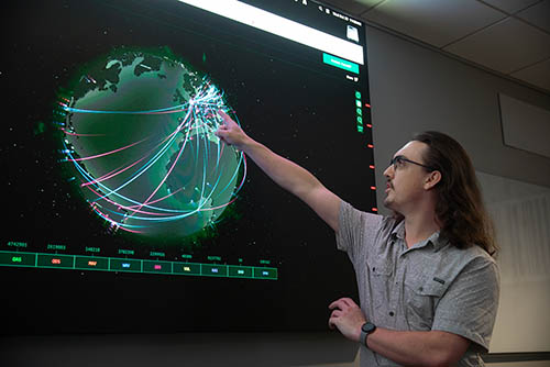 a cybersecurity major stands at the large screen on the wall featuring a graphic of the globe and points to the screen