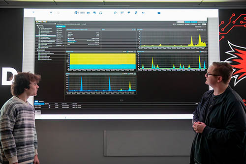 two cybersecurity students stand at the large screen in the command center and study a graph