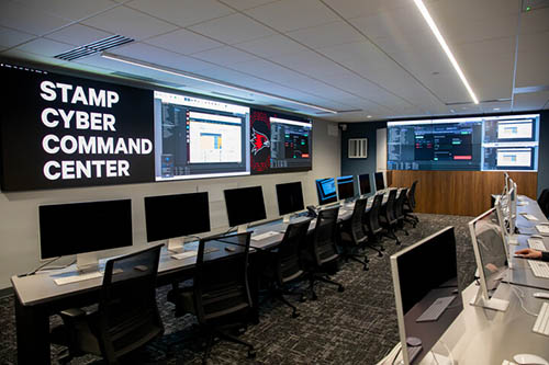 a wide view of the cyber command center featuring the wall screens and rows of computers 