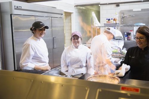 Hospitality management students smile while working next to a professor at a kitchen stove in their internship
