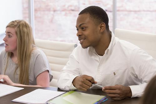 A student smiles while sitting at a table with peers in a management and marketing class.