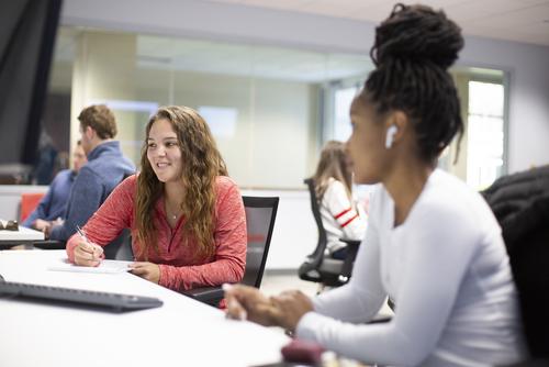 management major's in their internship expereincesmiles and writes in a notebook while working next to a peer in the sales lab.