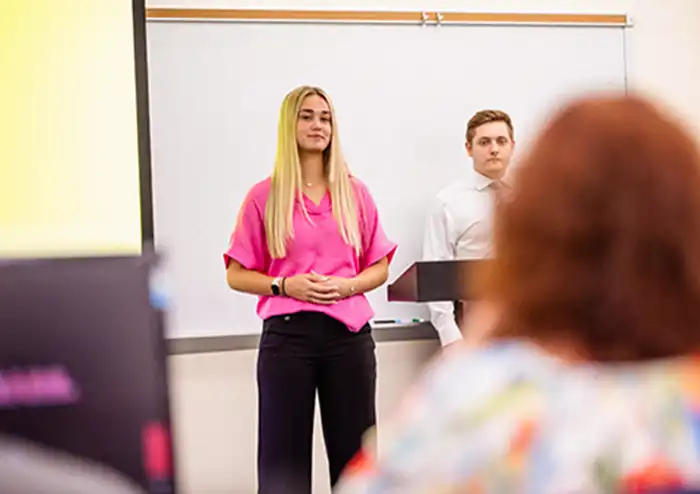 student in a pink dress shirt presents in front of a classroom showcasing her marketing experience and getting hands-on learning in presentation skills.   