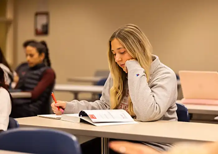 student in the marketing degree program sitting in class reading textbook and taking notes with pen and paper. 