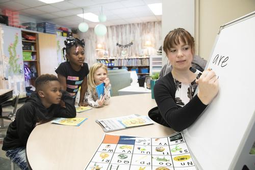 A student teacher writes on a dry erase board as students watch while sitting at a table.  