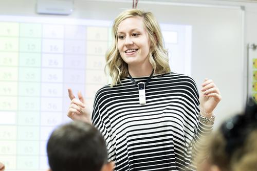A student teacher smiles while talking in front of a class of children at a local school.  