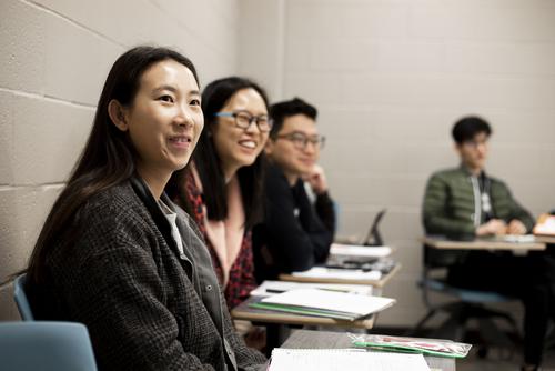 Students smile while sitting at desks and listening in a TESOL class. 