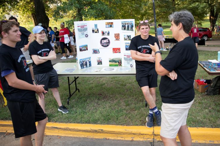 Three sports management students stand in front of their sports management society poster while conversing with a faculty memeber.