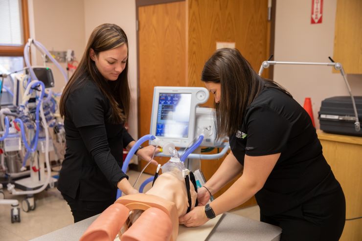 Two respiratory therapy studetns practice on a dummy while reading vital signs.