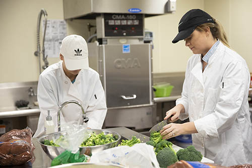 two bachelor of science in nutrition students in kitchen uniforms chop broccoli in an industrial kitchen