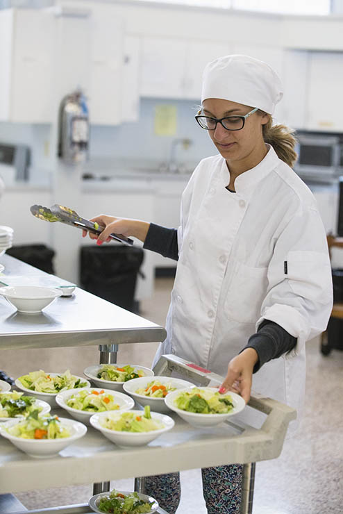 a nutritionist program student in fully professional kitchen garb works to make salad bowls and place them on a cart