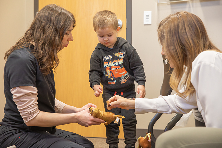 Twoa comm disorders students sit with a child patient and points at a T-Rex toy.