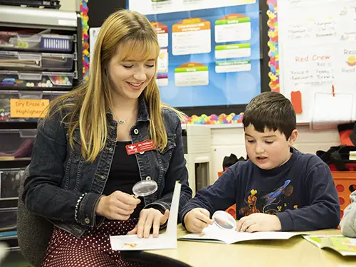 a student teacher from SEMO works in a classroom looking at a book using a magnifying glass next to a young boy doing the same