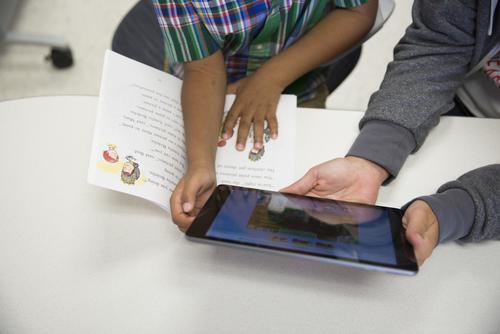 Hands of a young child grasp a book and a tablet as a teacher's hands also hold the tablet.