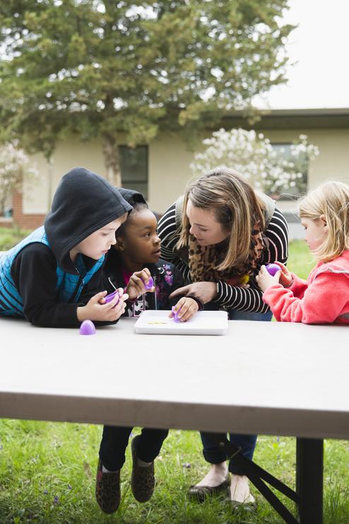 A student teacher leans over and instructs three children outside.