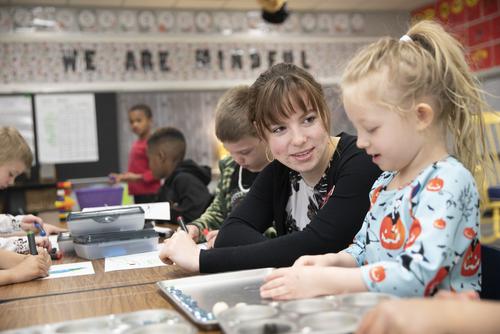 A student teacher wearing a black sweater works with a young girl wearing a blue dress in a classroom.