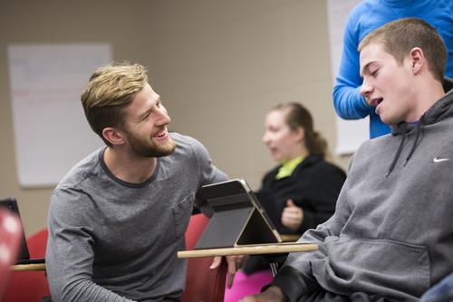  A student kneels down to have a conversation with another student sitting in a desk with their iPad. 